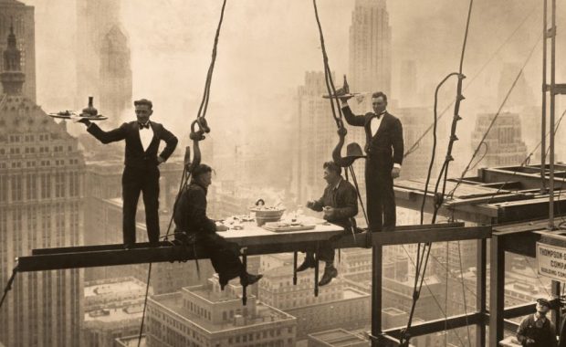 Construction workers sitting on beams suspended high above New York City while eating a gourmet meal.