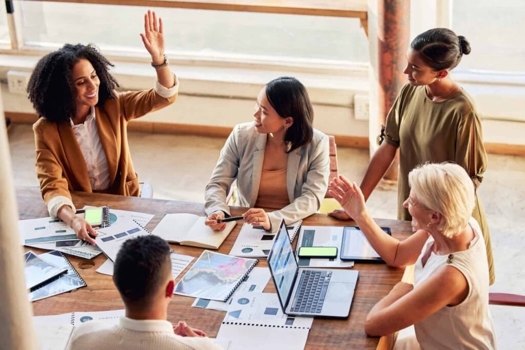 A diverse group of female professionals high-fiving, personalized care and collaborating around a wooden table with laptops and documents.
