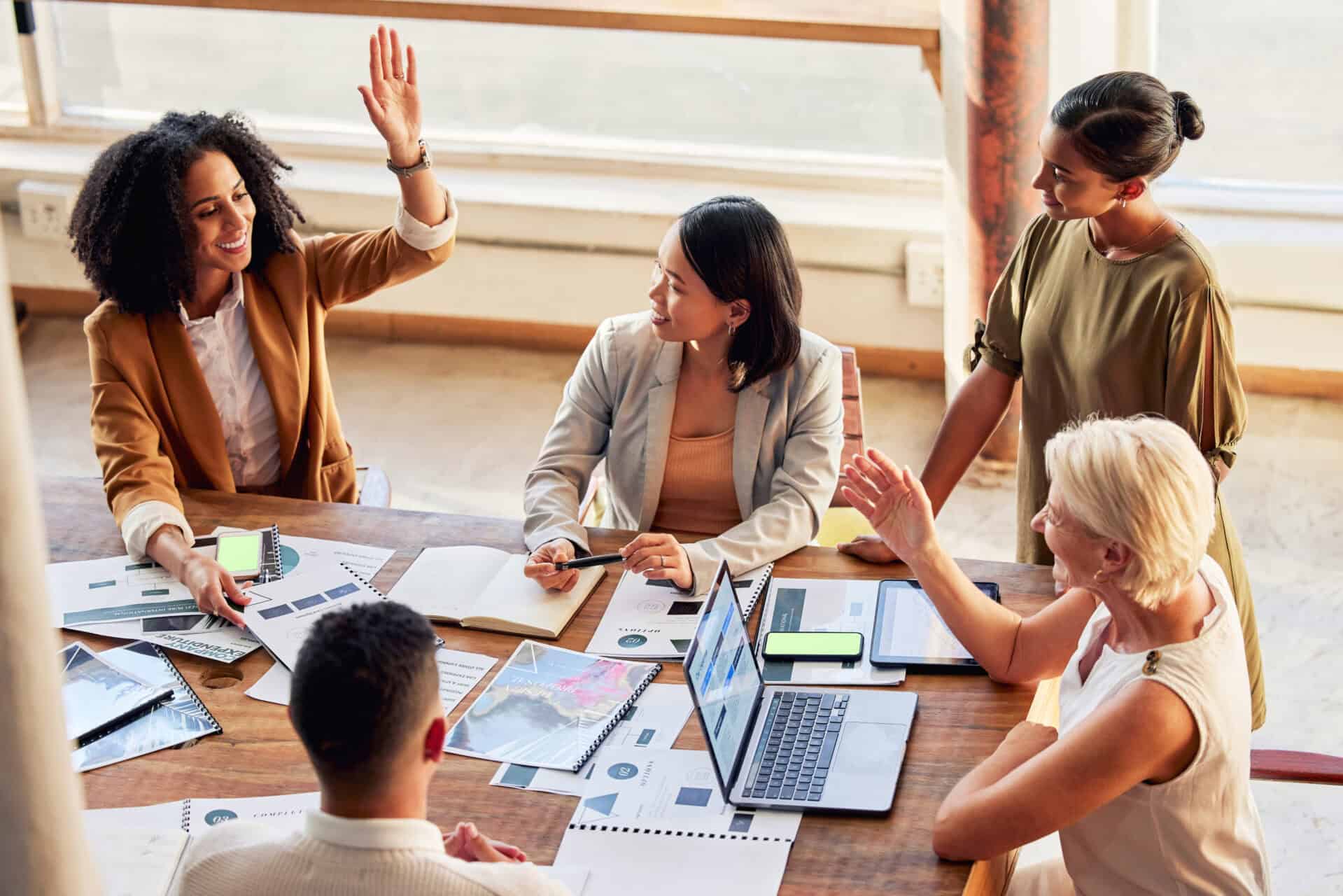 A diverse group of female professionals high-fiving and collaborating around a wooden table with laptops and documents.