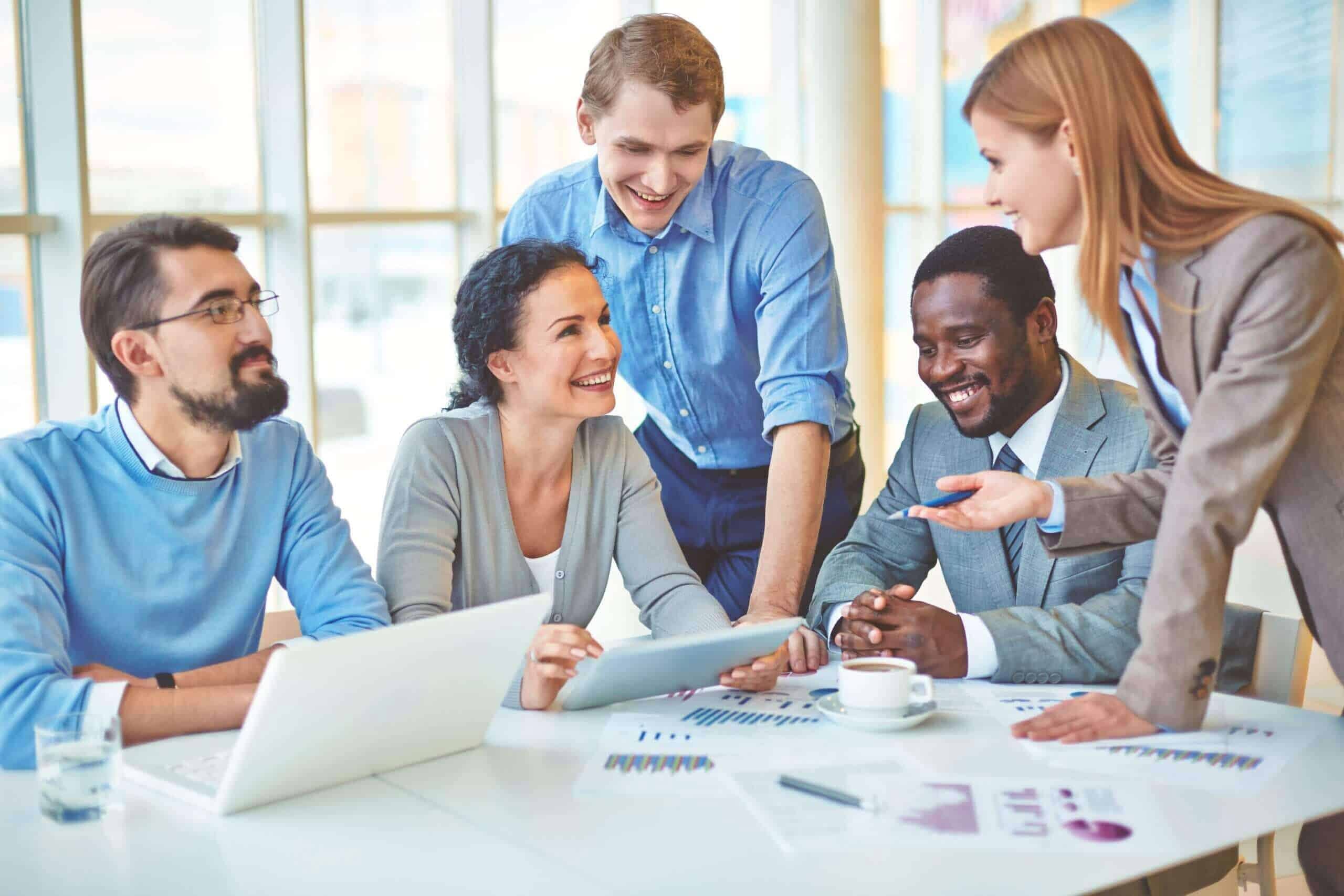 A diverse group of smiling professionals in a modern office setting discussing documents and a tablet during a strategy session.