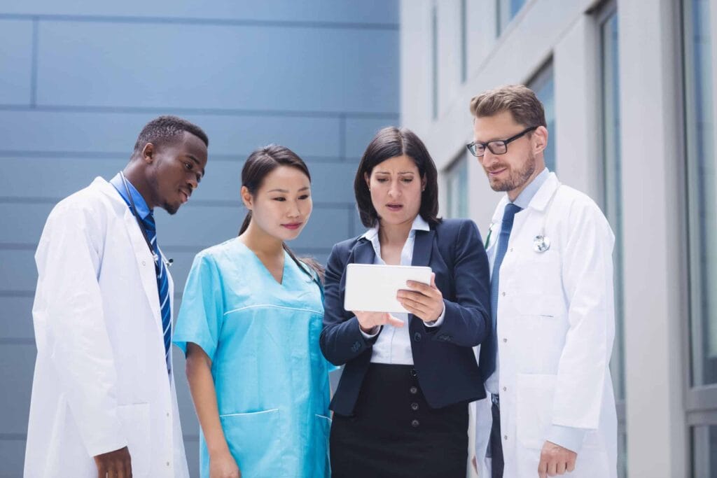 A diverse group of doctors and a nurse in lab coats and scrubs looking at a digital tablet held by a woman in a business suit.