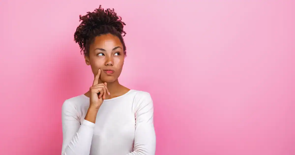 A young woman with curly hair tied up, looking thoughtful with her finger on her chin against a solid pink background with ample copy space and thinking about pulse surveys.
