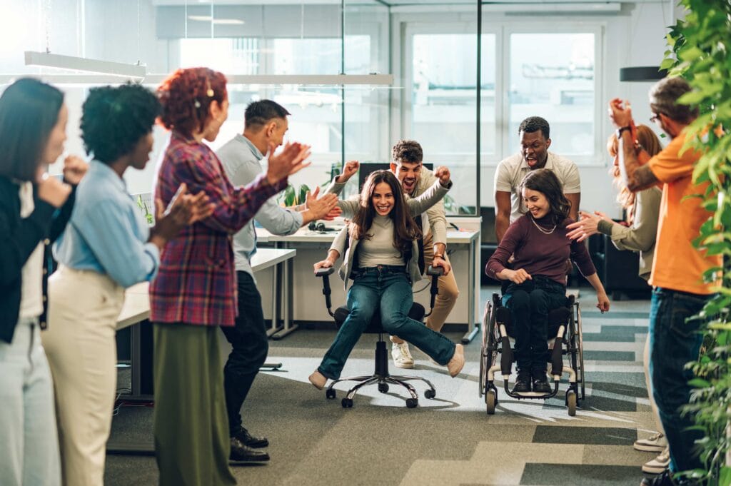 A diverse, multiracial business team cheering enthusiastically during a playful office chair race. One woman is being pushed in a rolling desk chair while another team member in a wheelchair participates, showcasing an inclusive, high-energy, and fun corporate culture with employee engagement trends for 2026.
