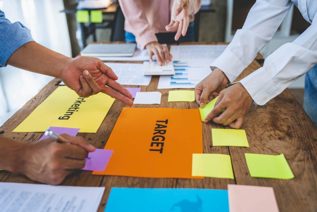 A close-up, high-angle view of business professionals' hands working around a wooden table with colorful sticky notes, a calculator, and papers labeled 