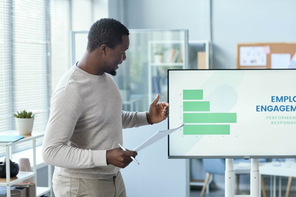 A professional Black man standing in a bright office, giving a presentation on employee engagement with a digital screen displaying a green bar chart titled Employee Engagement.
