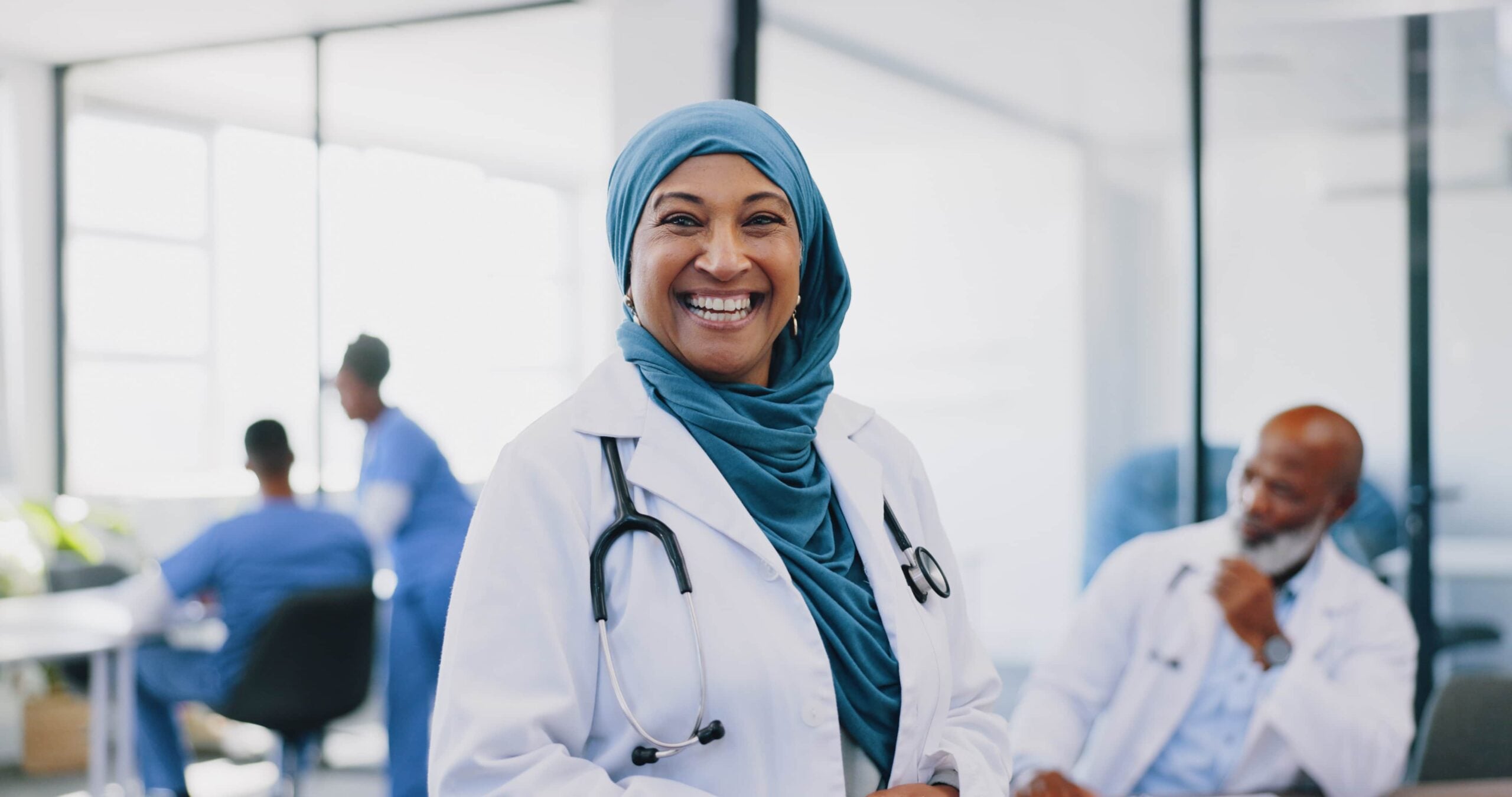 A smiling female doctor wearing a blue hijab and a white lab coat with a stethoscope around her neck, standing in a bright, modern hospital office with diverse colleagues in the background. Cultivating engagement.