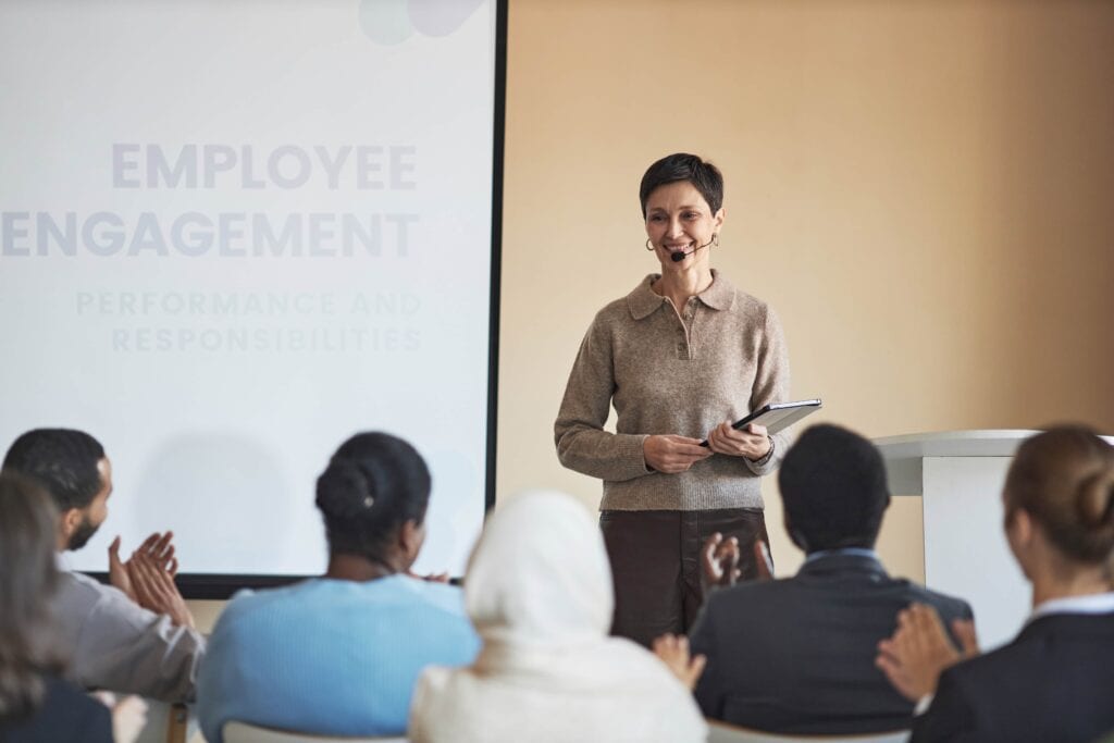 A smiling female professional speaker wearing a headset microphone and holding a tablet, standing in front of a presentation screen titled 