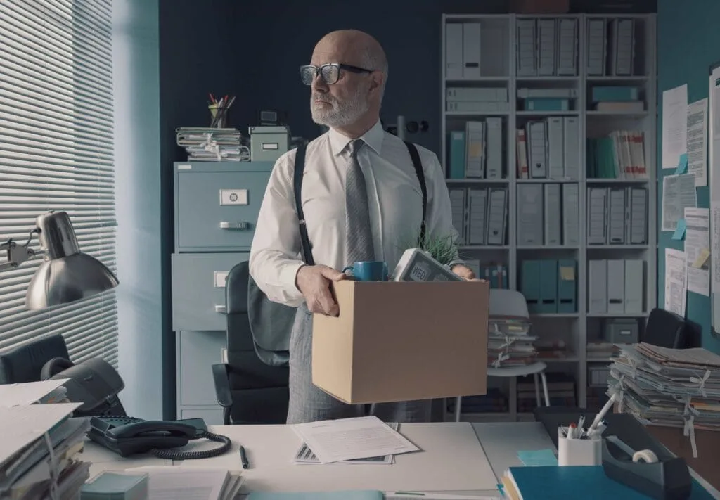 A senior businessman in a shirt and suspenders holding a cardboard box of personal belongings while leaving his office, representing career transition services in Chicago, Illinois.
