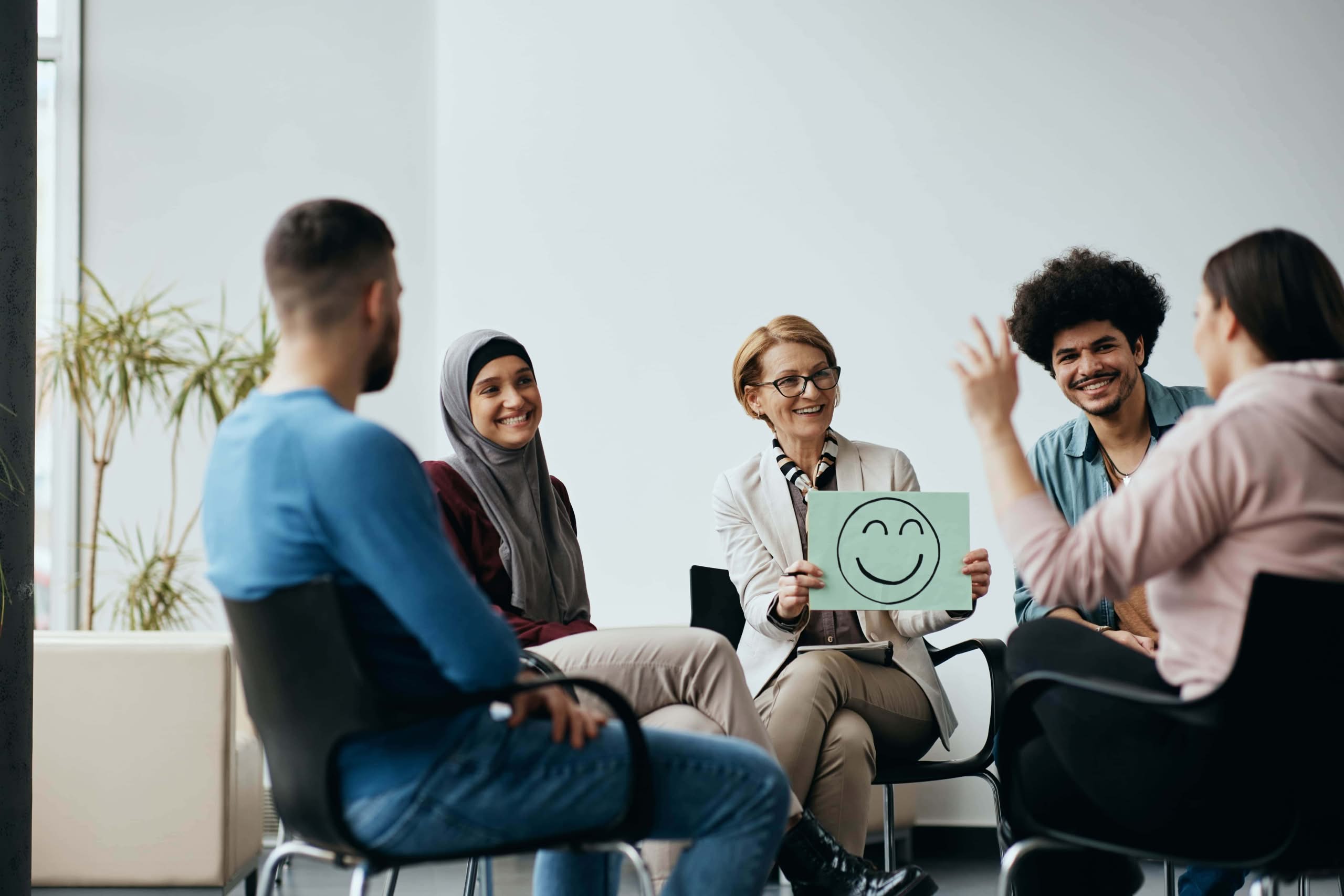 A smiling female therapist holding a green placard with a smiley face during a diverse group therapy session with young adults sitting in a circle. corporate mental health support initiatives