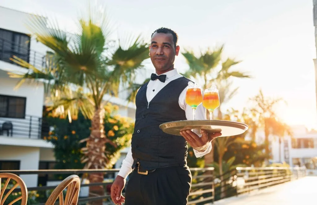 A professional hotel waiter in a formal vest and bowtie serving vibrant cocktails on a sun-drenched outdoor patio, providing elite guest service in Miami Beach, Florida.