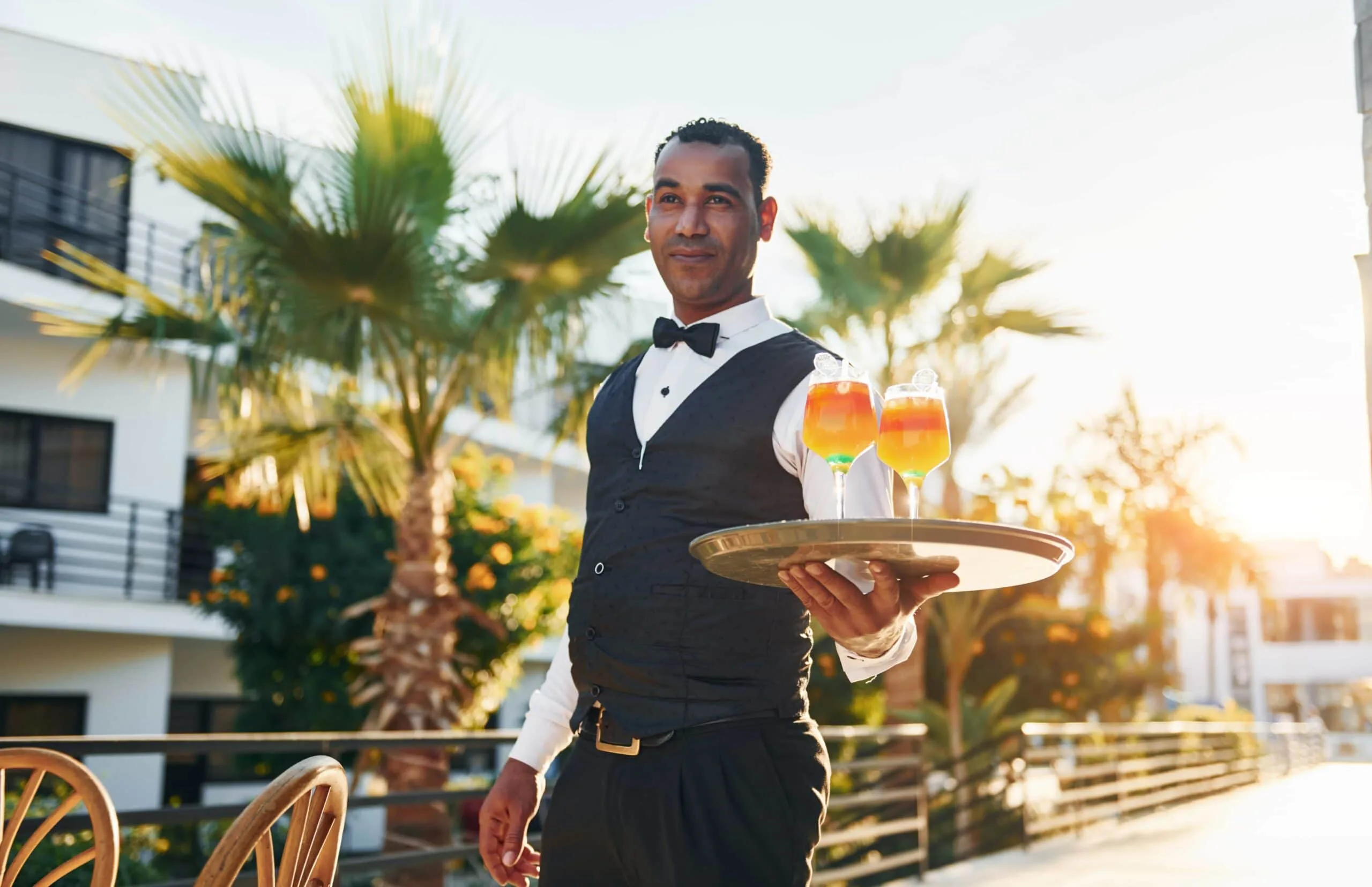 A professional hotel waiter in a formal vest and bowtie serving vibrant cocktails on a sun-drenched outdoor patio, providing elite guest service in Miami Beach, Florida. beating the summer slump