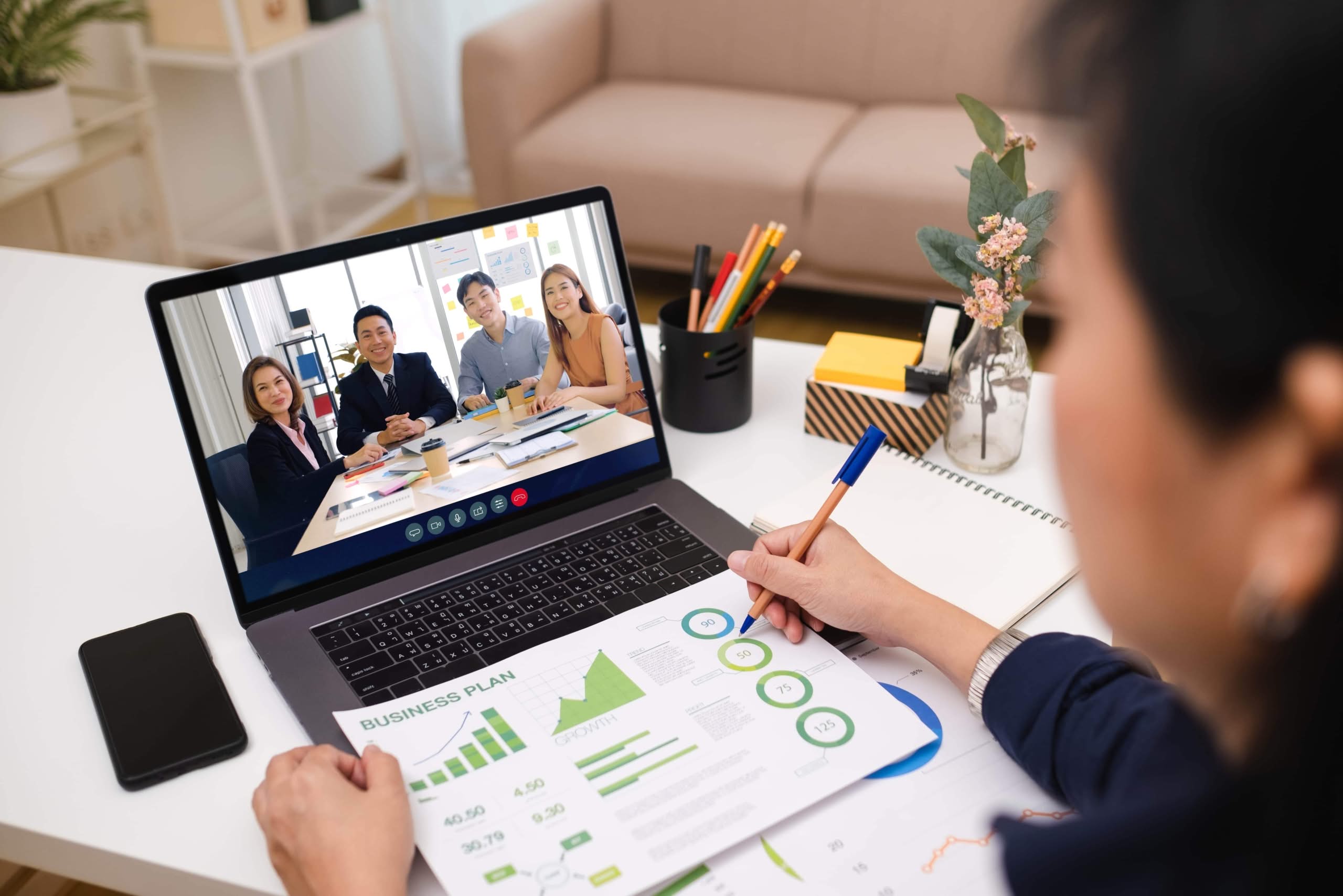 An over-the-shoulder view of an Asian woman working from home, presenting a business plan with green charts during a video conference call with colleagues on a laptop. Remote employee engagement survey