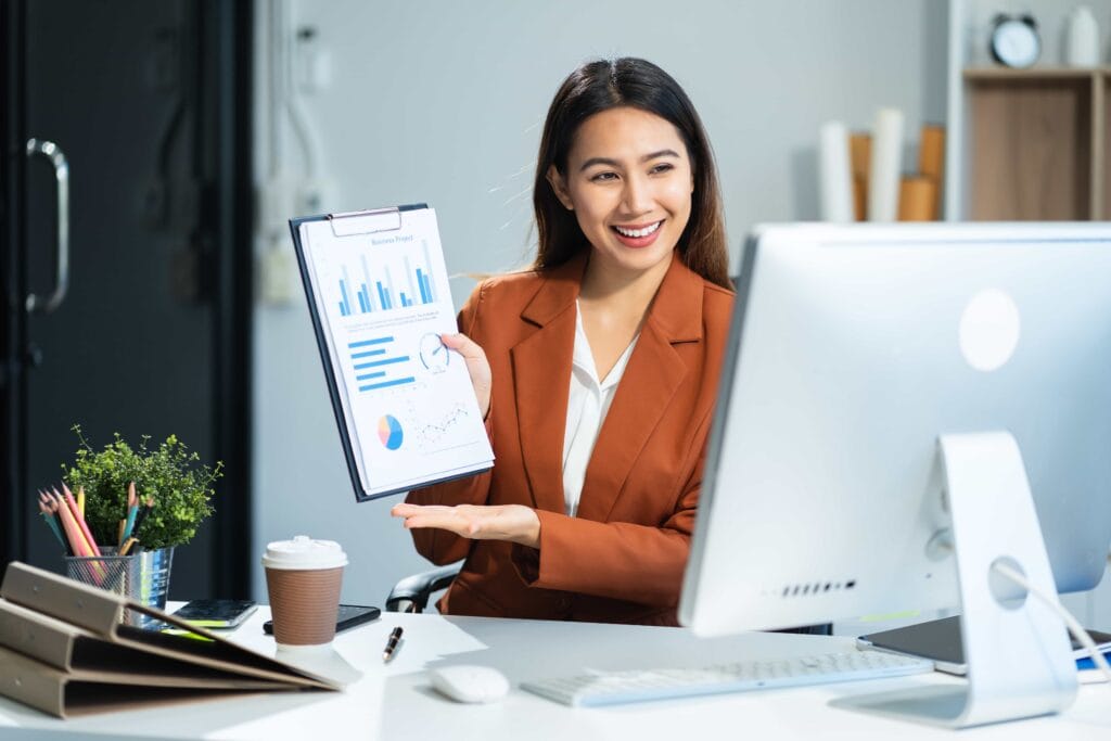 A smiling female professional in a brown blazer presenting a data report with charts and graphs at her desk, showcasing expert business consulting services in Manila, Philippines.