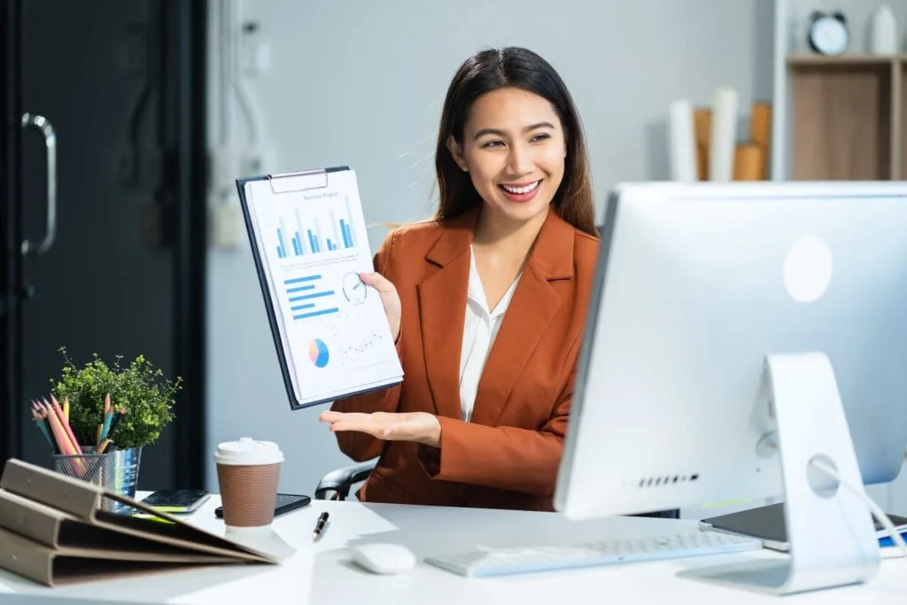 A smiling female professional in a brown blazer presenting a data report with charts and graphs at her desk, showcasing expert business consulting services in Manila, Philippines.