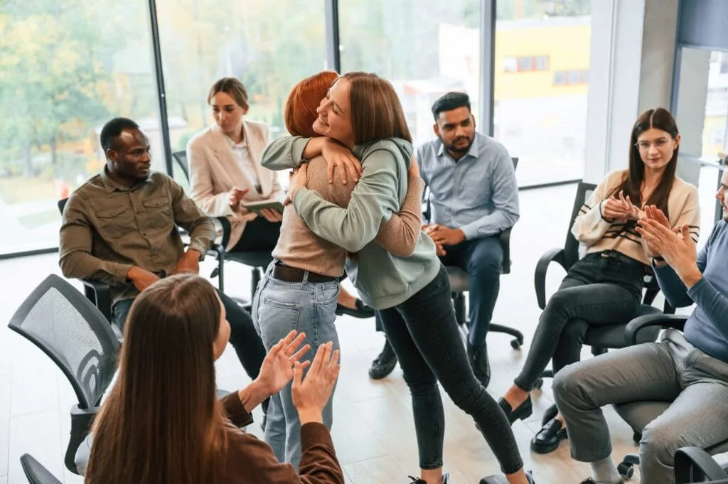 A diverse group of people sitting in a circle in a bright office, clapping as two women share a celebratory hug, highlighting a successful mental health support group in Ocala, Florida.