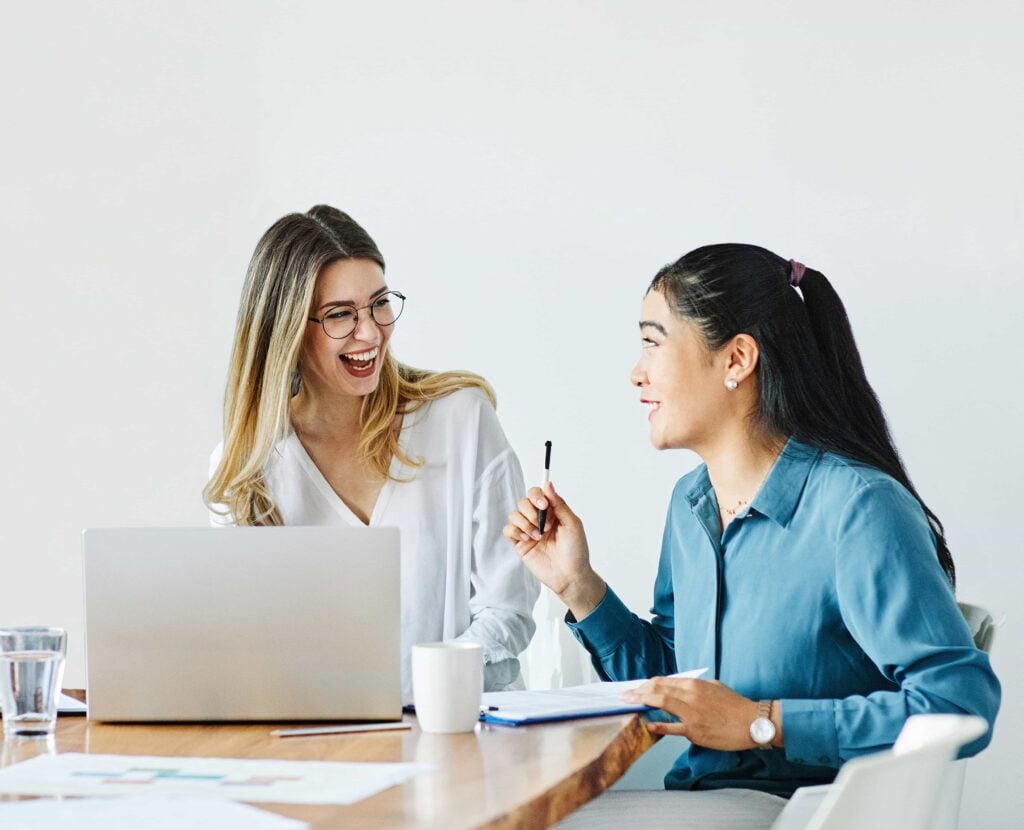 Two professional women collaborating and laughing during a business meeting with a laptop and documents on a desk in Denver, CO.