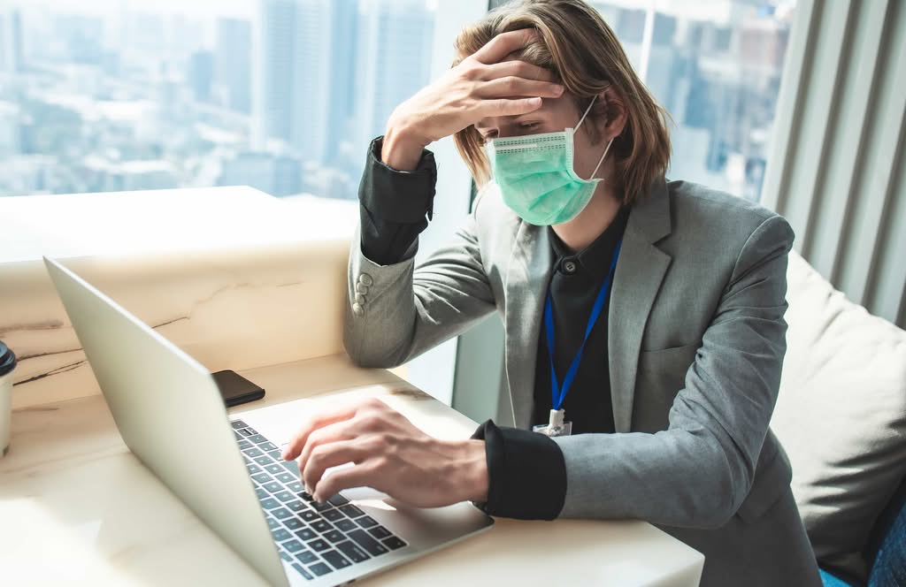 A stressed hospitality employee wearing a face mask and lanyard sitting at a desk indoors while working on a laptop with a hand on his forehead in Orlando, FL.