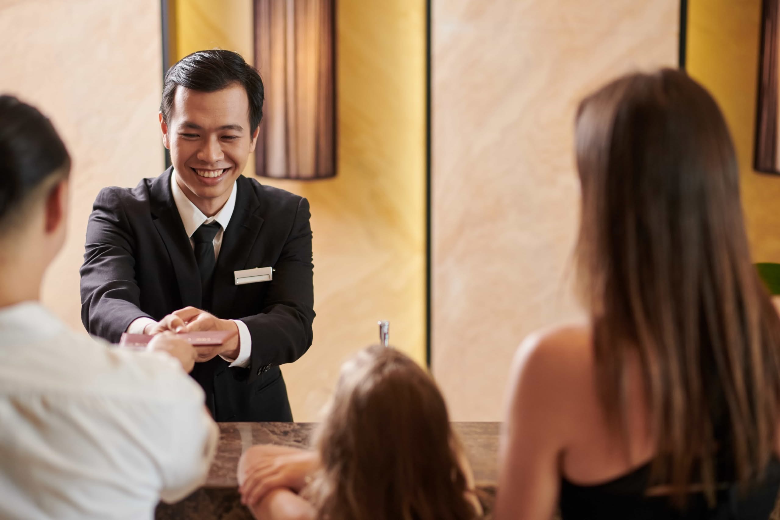 A smiling male hotel receptionist in a black suit helps a family check in at a luxury front desk, demonstrating effective hotel staff retention strategies in Las Vegas, NV.