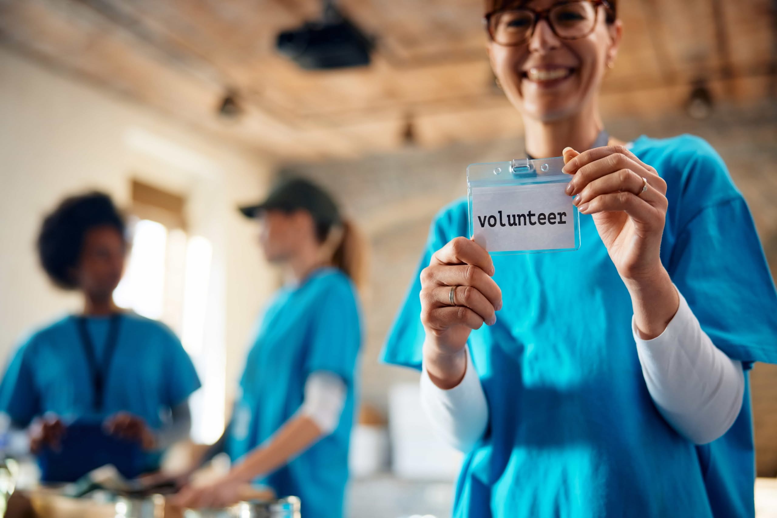 A smiling woman in a blue t-shirt holds up her clear volunteer identification badge while other employee retention strategies for volunteers work in the background of a nonprofit organization in Austin, TX.